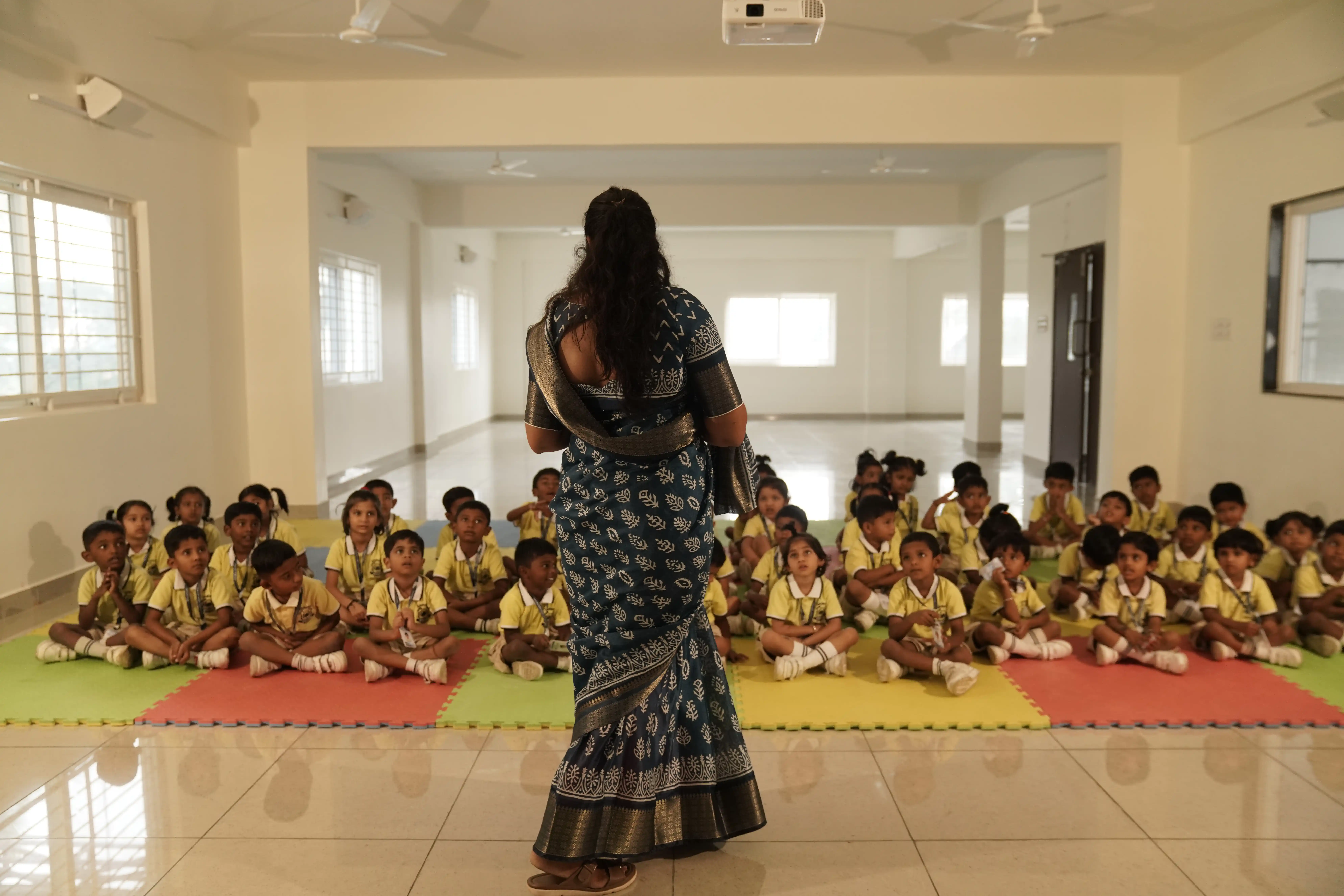 Teacher leading pre-school children seated on play mats in the activity hall at Takshashila School